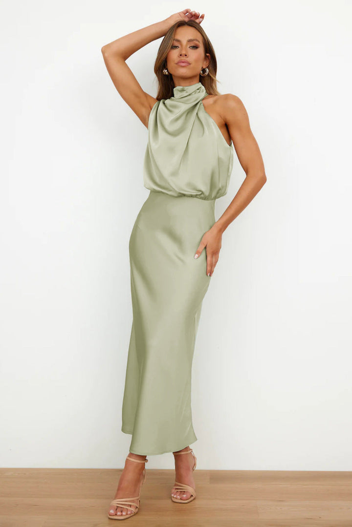 Sage green sleeveless halter dress with elegant draping in a minimal indoor setting.