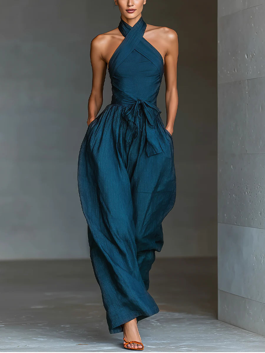Elegant blue halter-top jumpsuit with flowing wide legs, styled with orange sandals in a minimalist indoor setting.