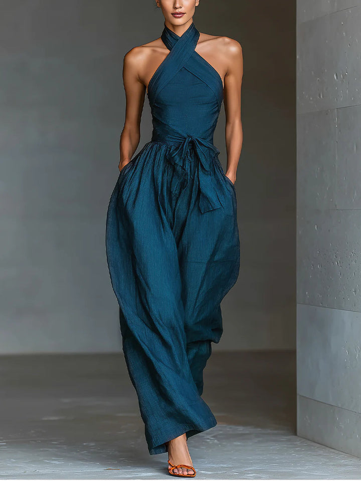 Elegant blue halter-top jumpsuit with flowing wide legs, styled with orange sandals in a minimalist indoor setting.