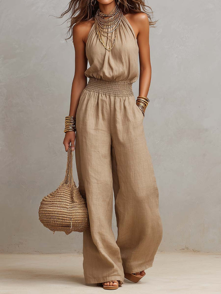 Elegant jumpsuit in beige with halter neck and smocked waist, accessorized with layered necklaces and bracelets. Studio setting.