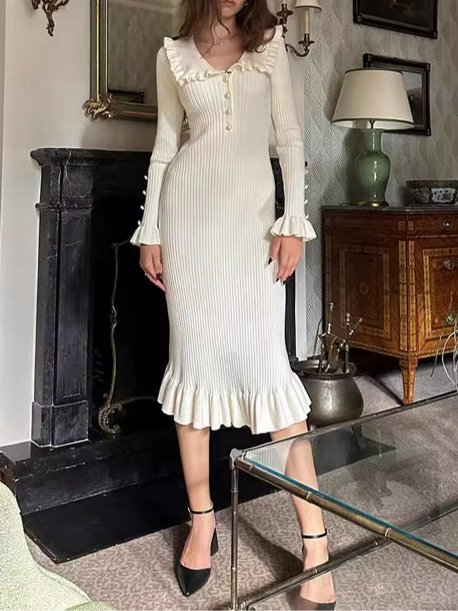 Model in elegant cream knit dress with ruffled collar and cuffs, pearl buttons, standing in a vintage decorated room with a fireplace.