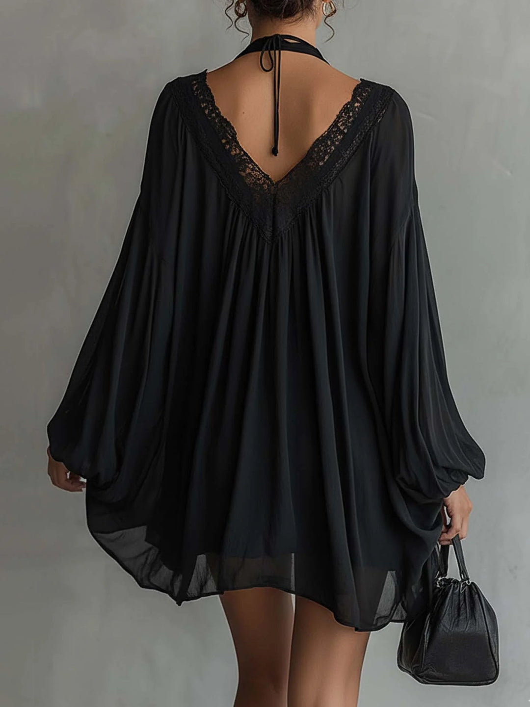 Back view of black mini dress with lace detailing and flowing sleeves, paired with a black bag, set against a gray studio background.