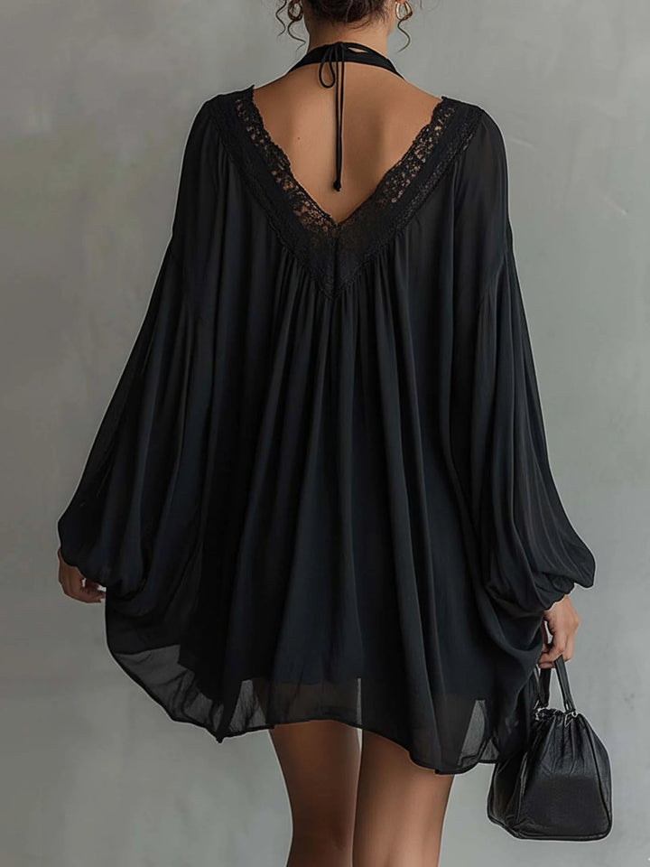 Back view of black mini dress with lace detailing and flowing sleeves, paired with a black bag, set against a gray studio background.