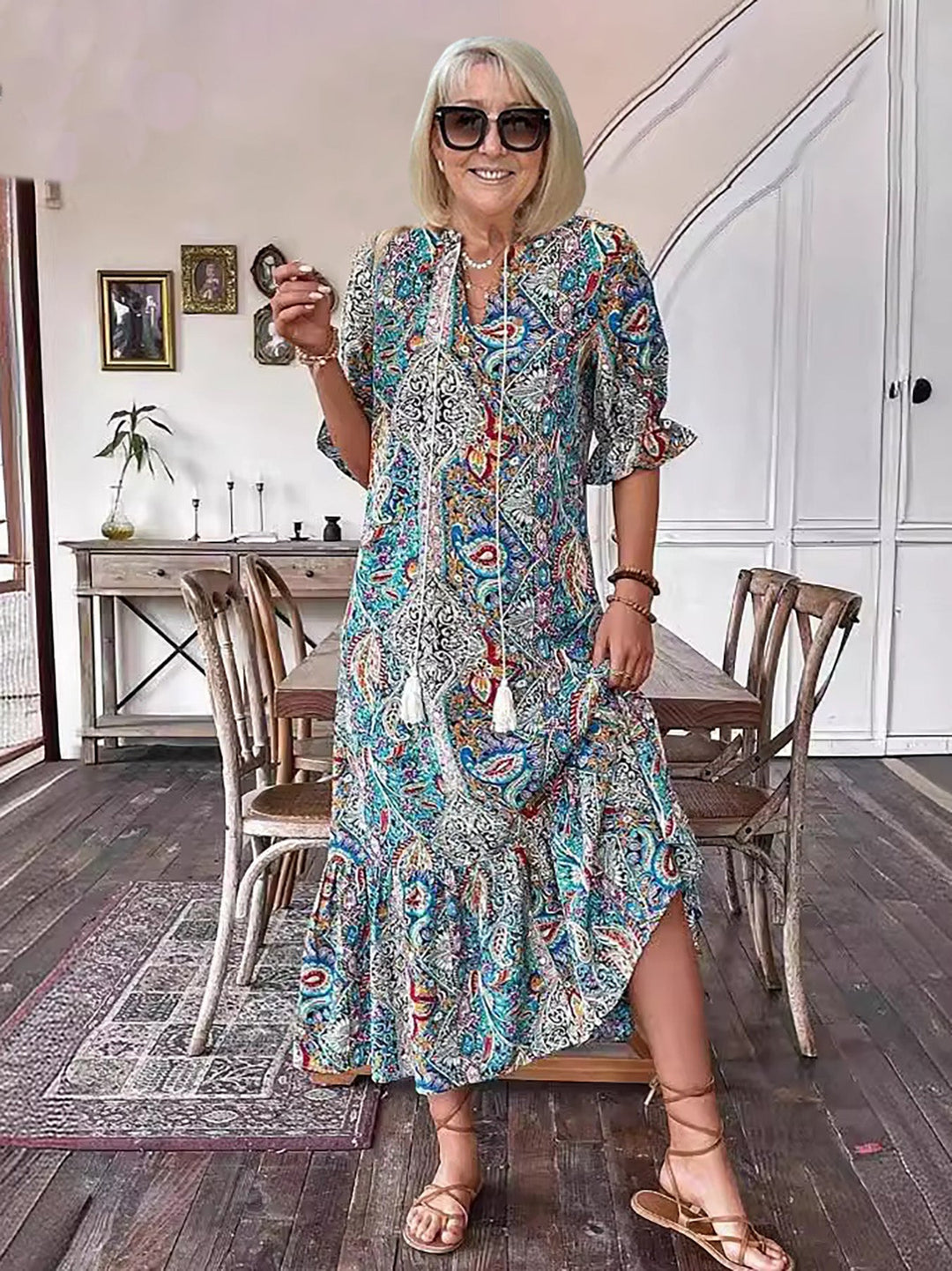 Woman in boho dress with intricate patterns, smiling indoors, wooden furniture, standing casually, bright ambiance.