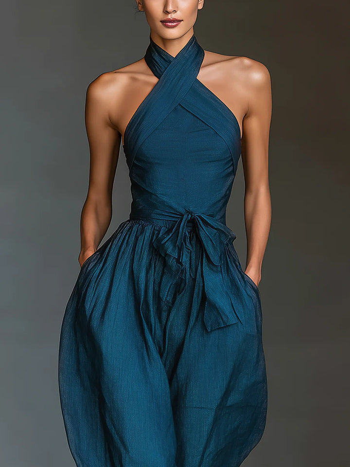 Front close-up of blue halter-top jumpsuit with tied waist and wide legs in a minimalist indoor setting.