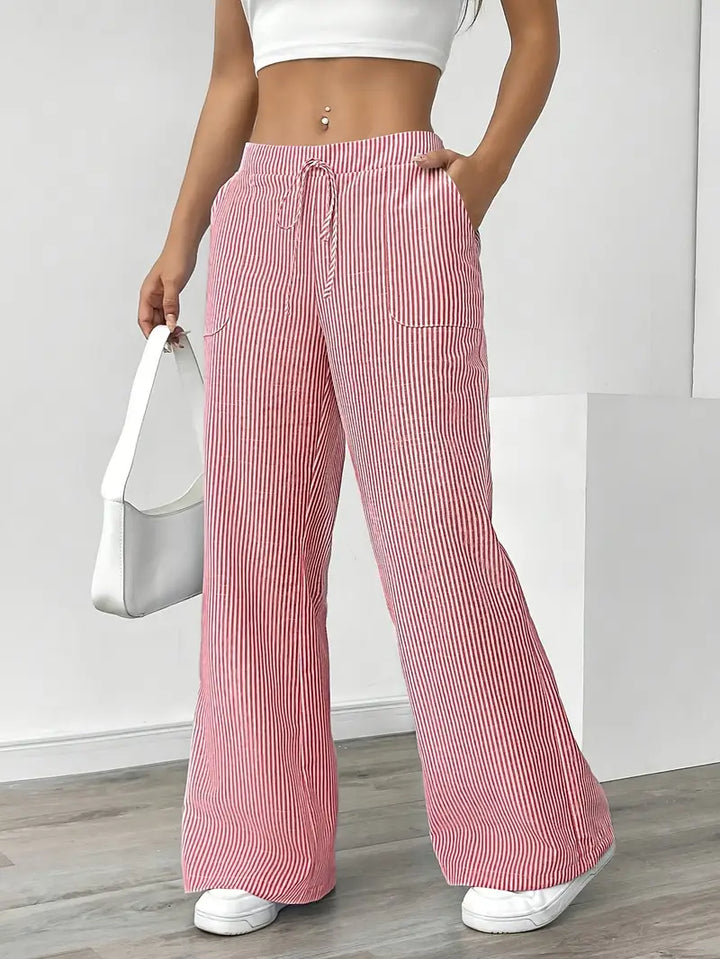 Red and white striped pants with pockets and drawstring, modeled indoors, paired with white sneakers and white bag.
