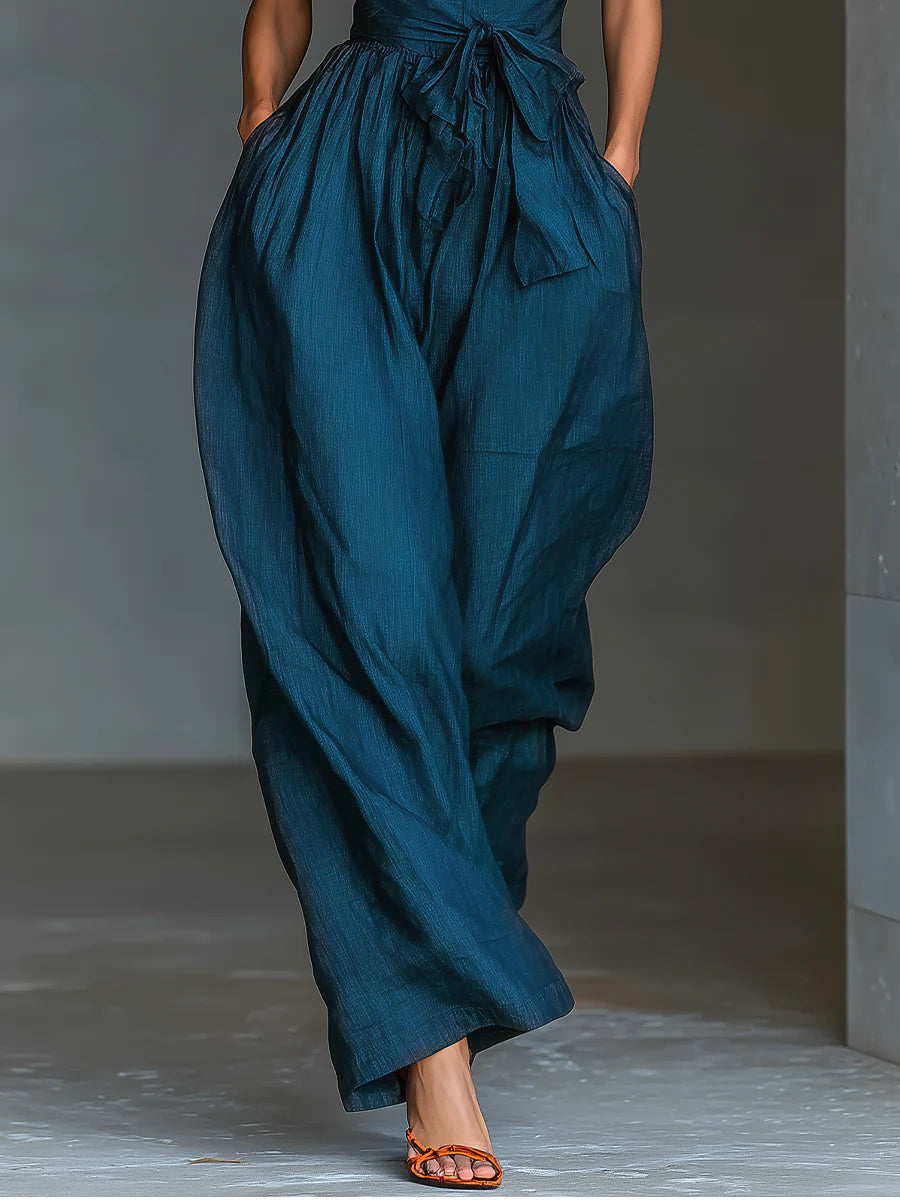 Blue wide-leg halter-top jumpsuit with tied waist, styled with orange sandals in a minimalist indoor setting.