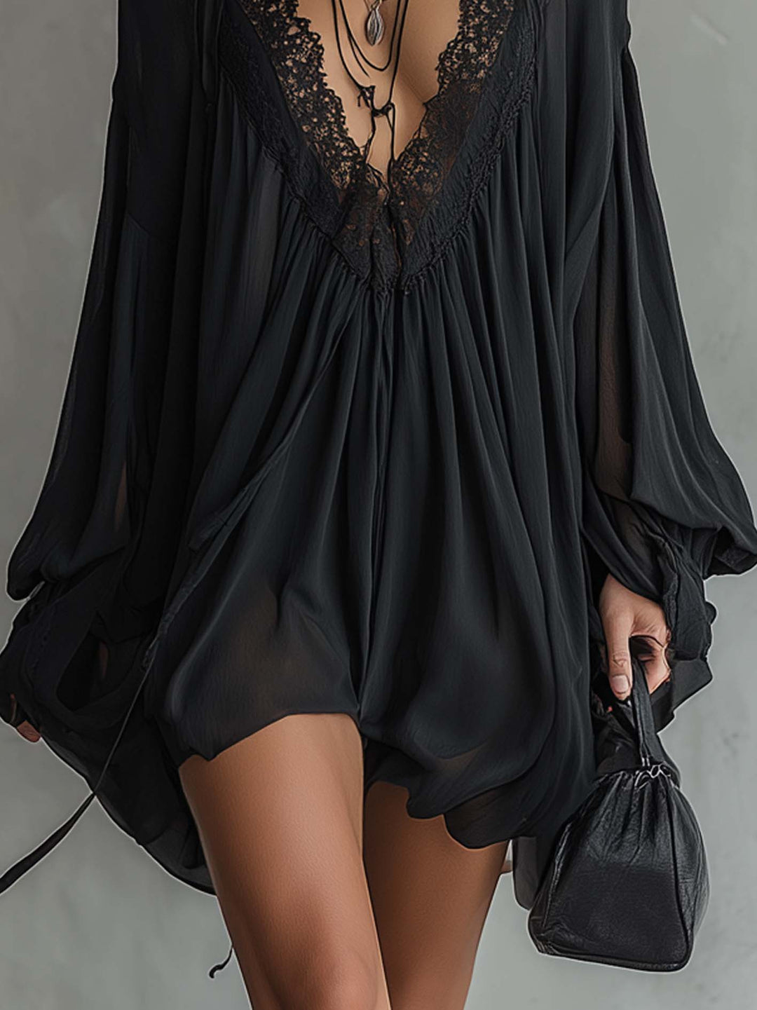 Close-up of black mini dress with lace neckline and flowing silhouette, holding a black bag, in a gray studio setting.