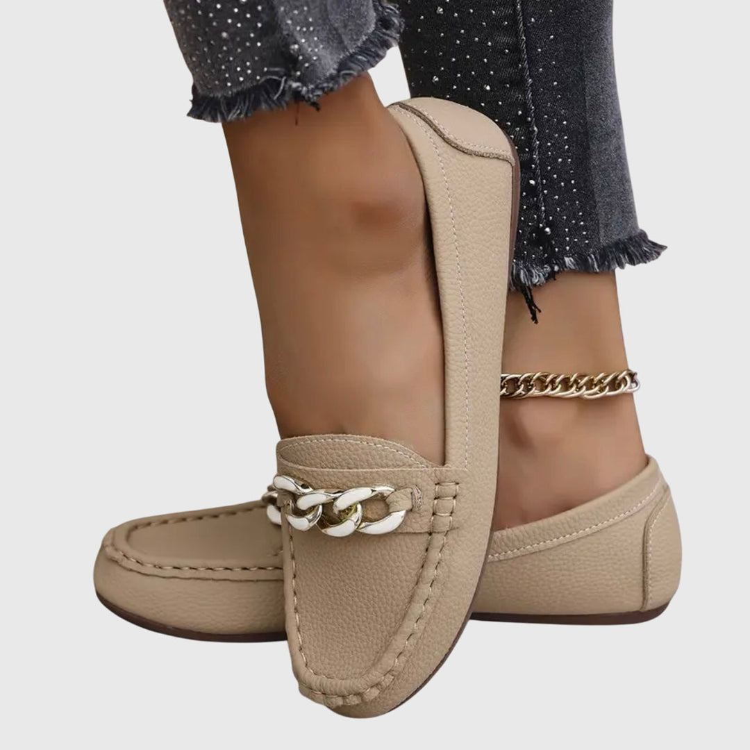 Side view of beige orthopedic loafers with chain detail, worn with dark pants on a gray background.