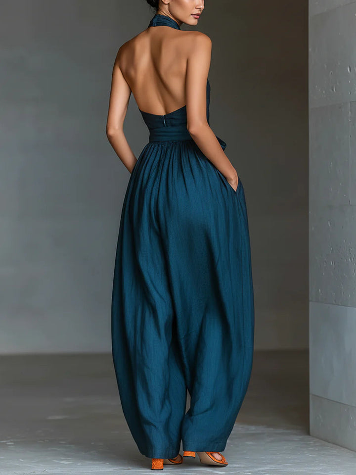 Back view of blue halter-top jumpsuit showing open back design, styled with orange sandals in a minimalist indoor setting.