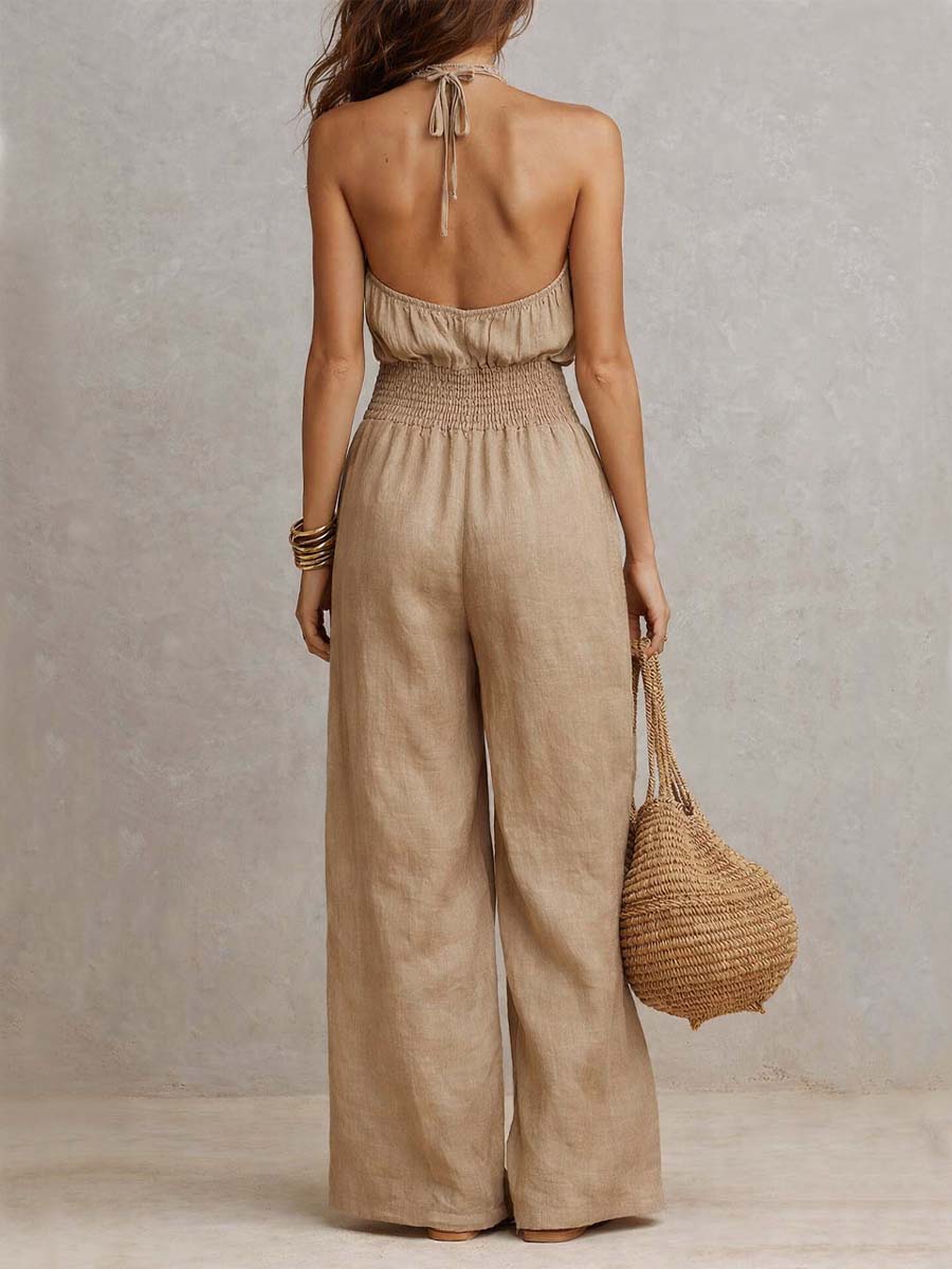 Back view of beige jumpsuit with tie-up halter neck and smocked waist, paired with a woven bag. Studio setting.