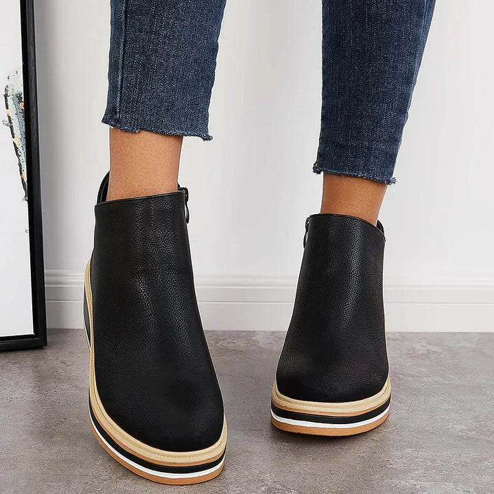 Front view of black luxe boots with wedge soles, worn with frayed jeans, on a gray floor.