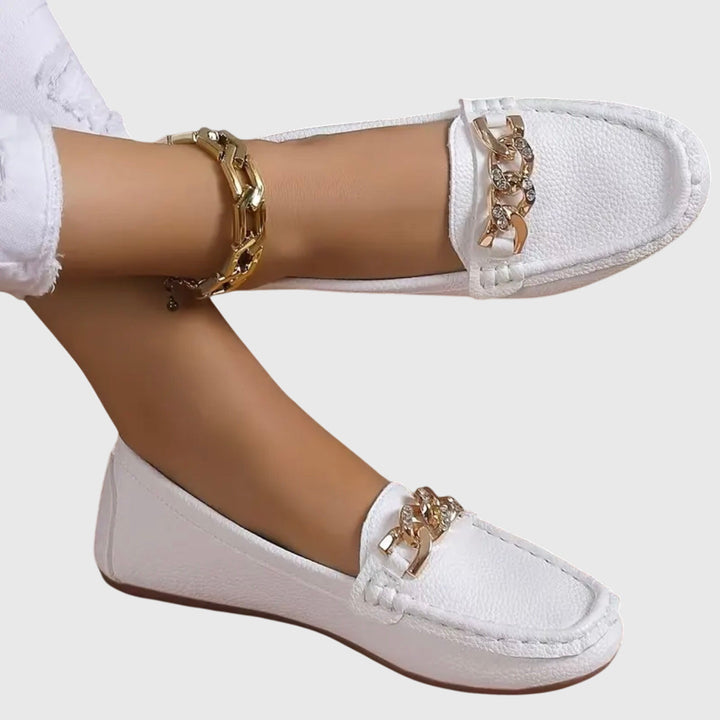 White orthopedic loafers with gold chain detail, worn with white pants on a gray background.