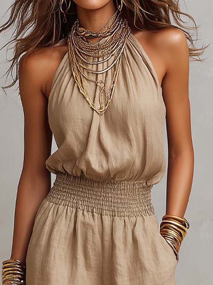Close-up of beige halter neck jumpsuit with elegant layered necklaces and smocked waist. Studio background.