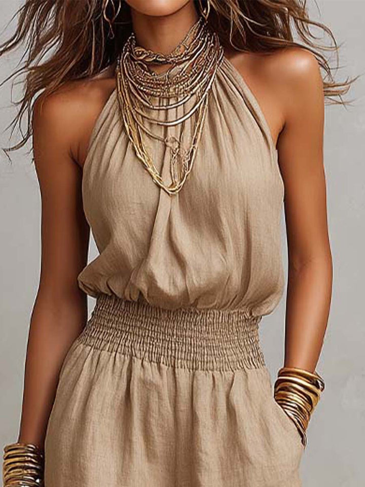Close-up of beige halter neck jumpsuit with elegant layered necklaces and smocked waist. Studio background.