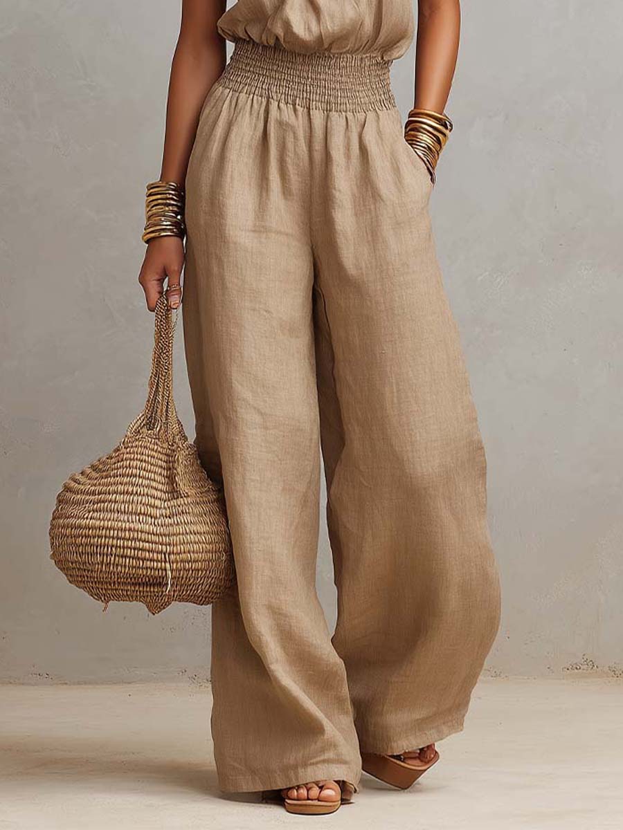 Beige jumpsuit featuring wide legs and smocked waist, accessorized with bracelets and woven bag. Studio scene.