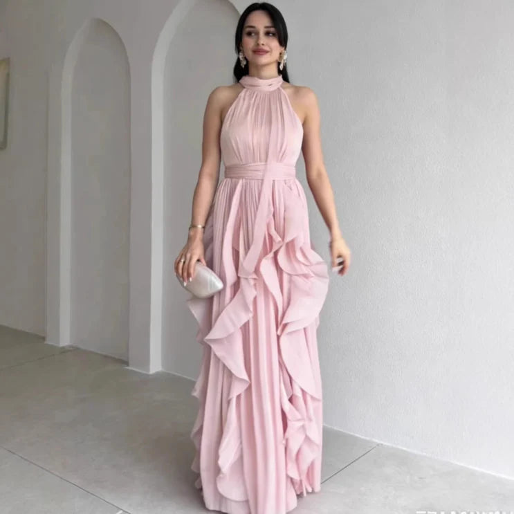 Model wearing a light pink high neck maxi dress with gathered ruffle layers, standing indoors against a white wall.