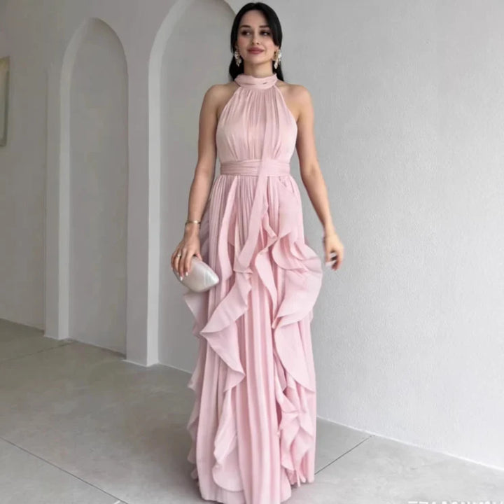 Model wearing a light pink high neck maxi dress with gathered ruffle layers, standing indoors against a white wall.