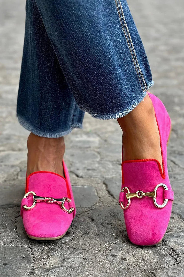 Bright pink loafers with metal detail, worn with frayed jeans on cobblestone street.