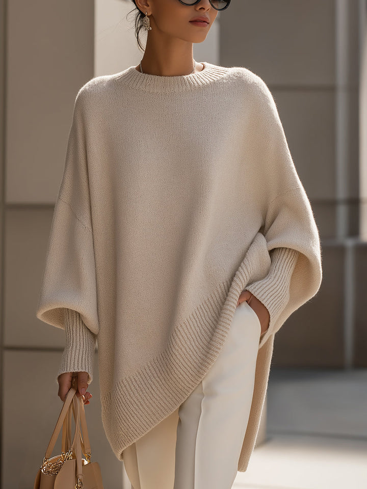 Woman in a loose beige sweater with ribbed hem and cuffs, wearing white pants and holding a bag, in an urban setting.
