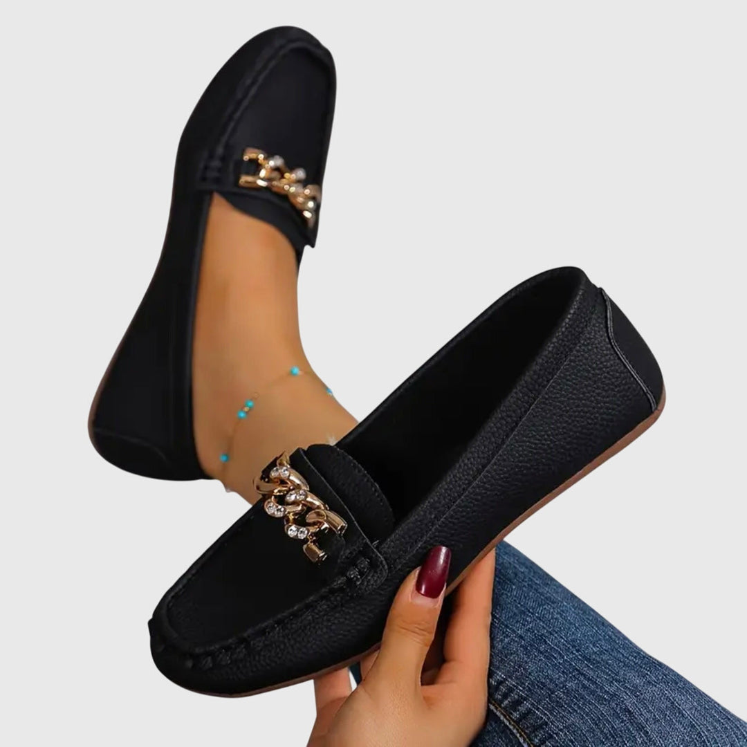 Black orthopedic loafers with gold chain detail, one held in hand, paired with blue jeans on a gray background.