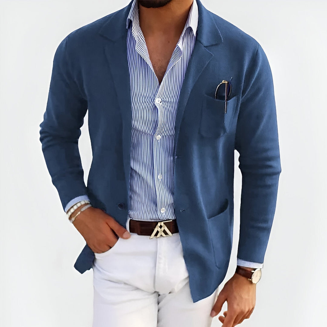 Man wearing a dark blue lightweight jacket over a striped shirt, paired with white pants, in a studio setting.