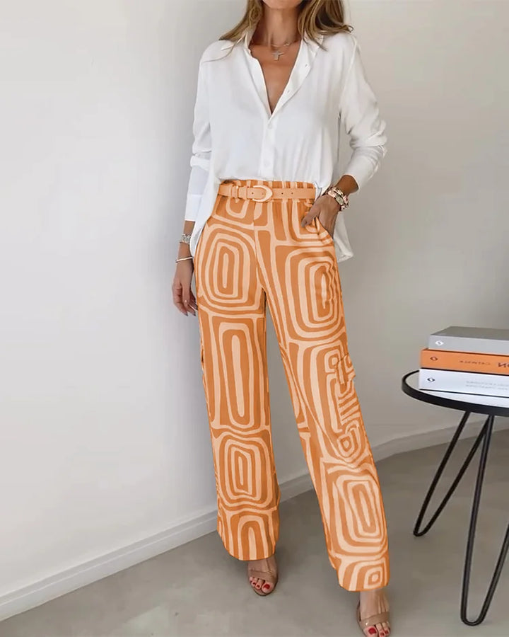 Elegant two-piece set featuring a white blouse and orange geometric-patterned pants. Displayed in a minimalist indoor setting.