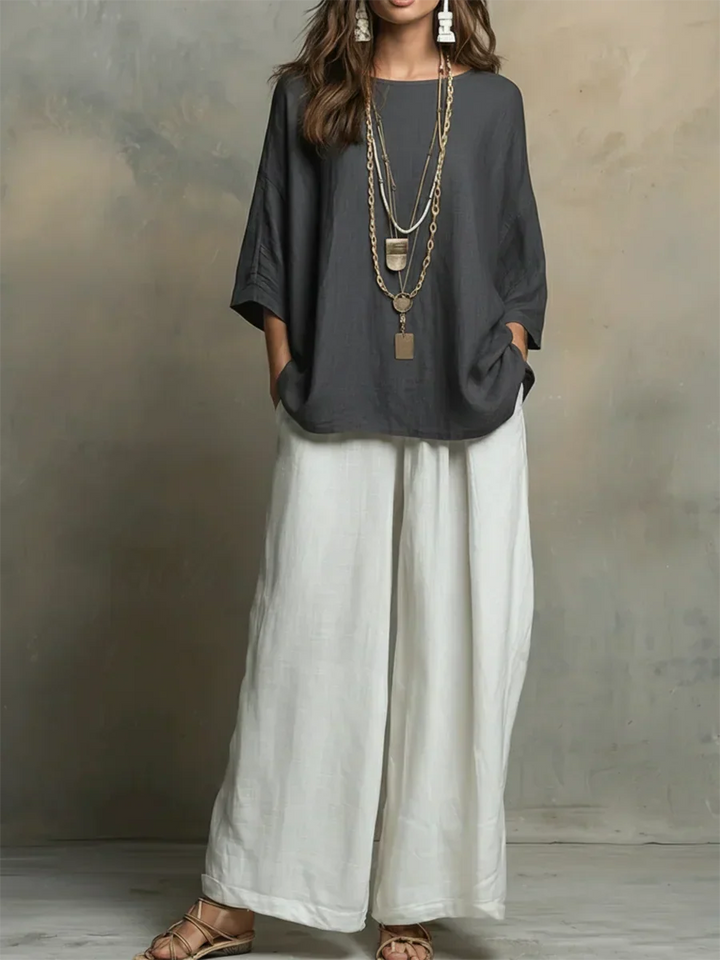 Woman in dark and white two-piece set, layered necklaces, hands in pockets, studio backdrop.