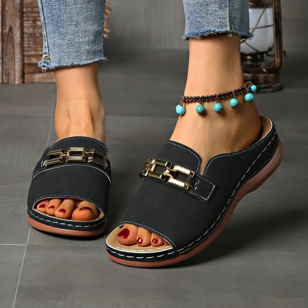 Black comfort sandals with gold chain accents on feet, worn with blue anklet, shown indoors on grey flooring.