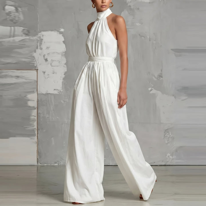 Model wearing a white halter-neck jumpsuit, standing sideways in a contemporary studio setting.