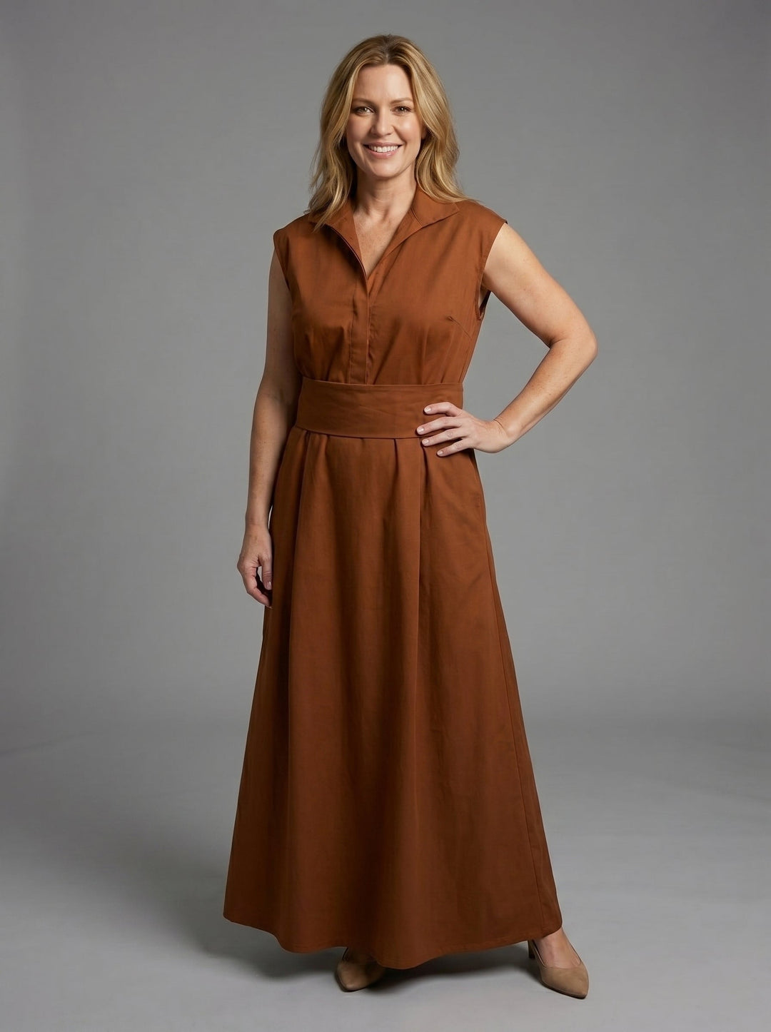 Smiling woman in brown sleeveless dress with wide waistband, posing in a studio with a gray background.