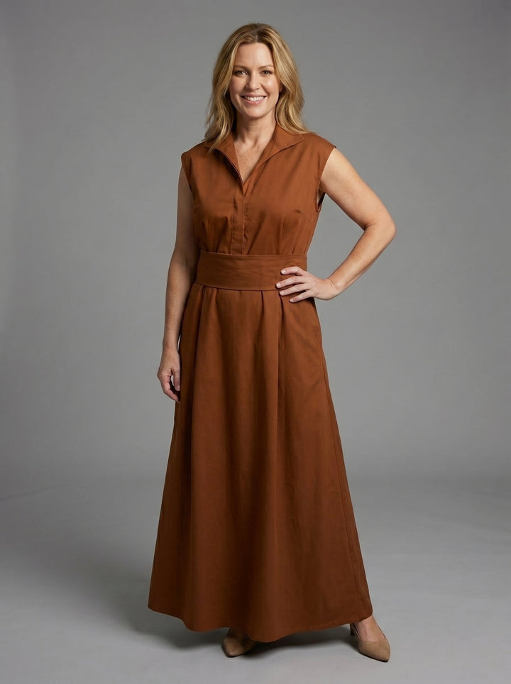 Smiling woman in brown sleeveless dress with wide waistband, posing in a studio with a gray background.