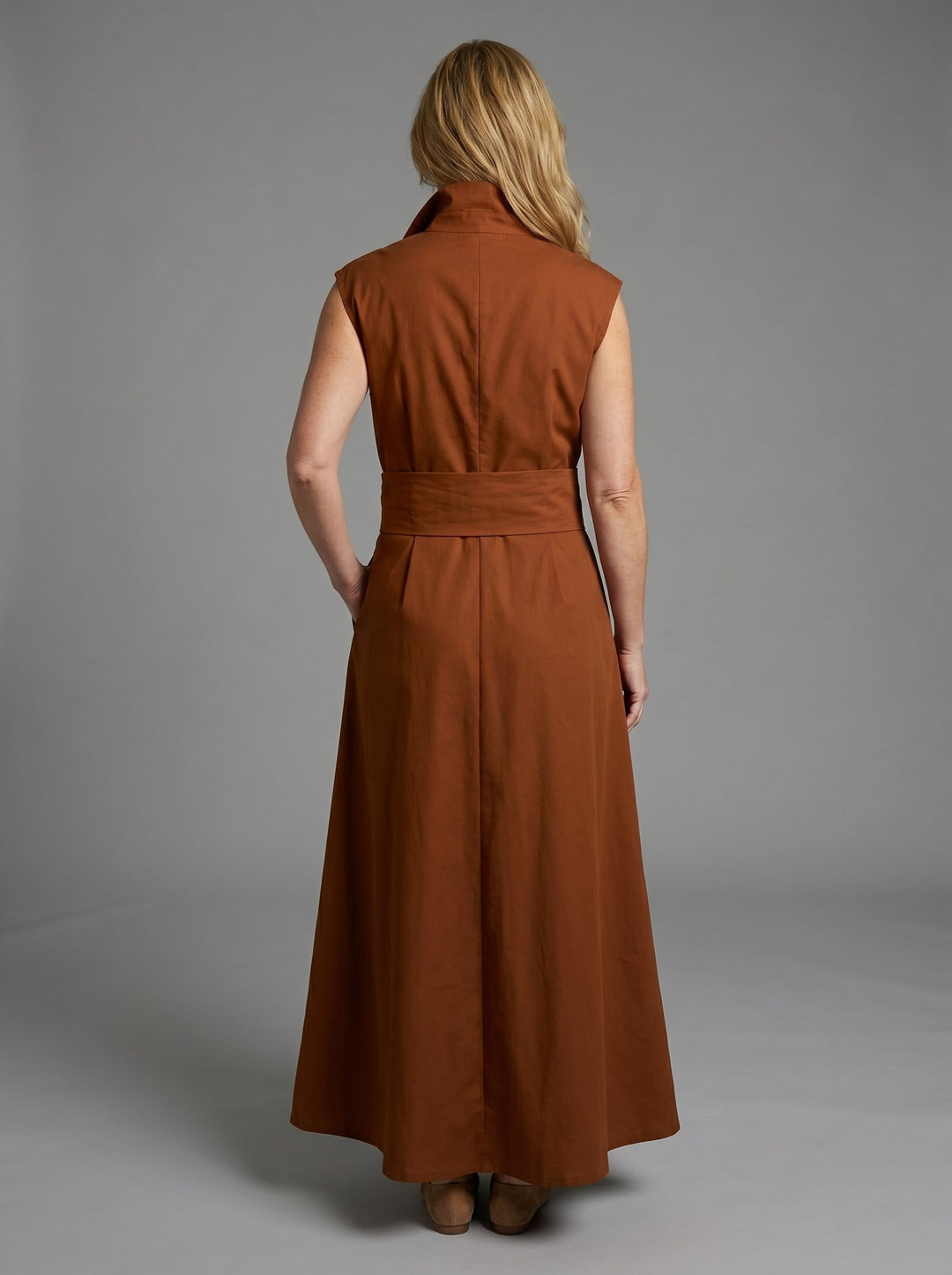 Back view of woman in brown sleeveless dress with wide waistband, posing in a studio with a gray background.