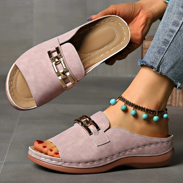 Pink comfort sandals with gold accents, held against indoor grey tiled background, worn with anklet.
