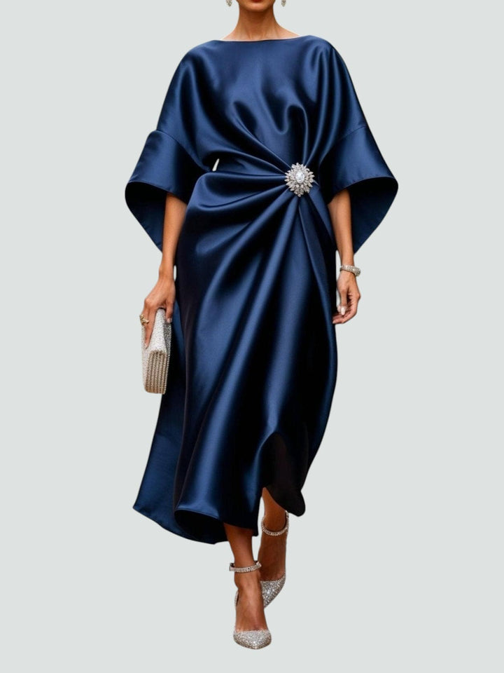 Elegant long satin dress in deep blue with a floral brooch, styled with sparkly heels and a matching clutch, studio background.