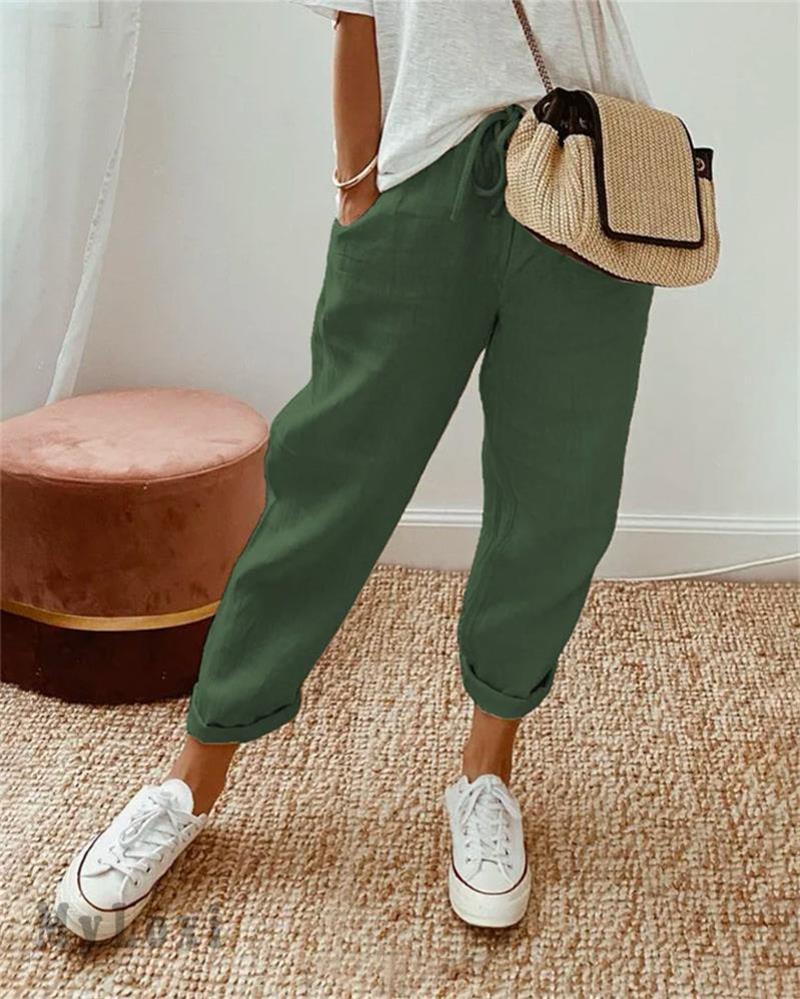 Green casual pants on model with white sneakers, white top, woven bag, indoor setting with rug and plush seat.