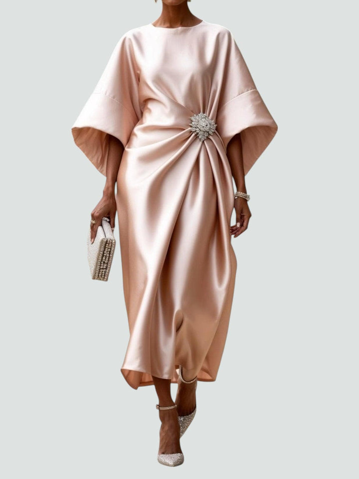 Elegant long satin dress in soft blush with a floral brooch, styled with sparkly heels and a matching clutch, studio background.