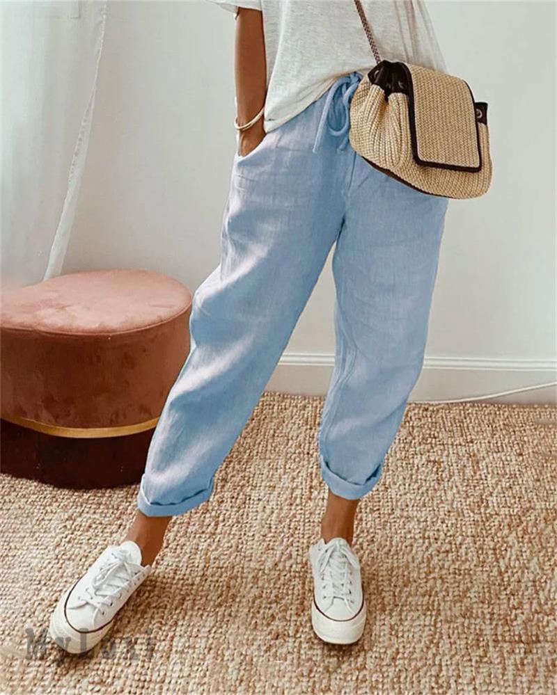 Light blue casual pants on model with white sneakers, white top, woven bag, indoor setting with rug and plush seat.