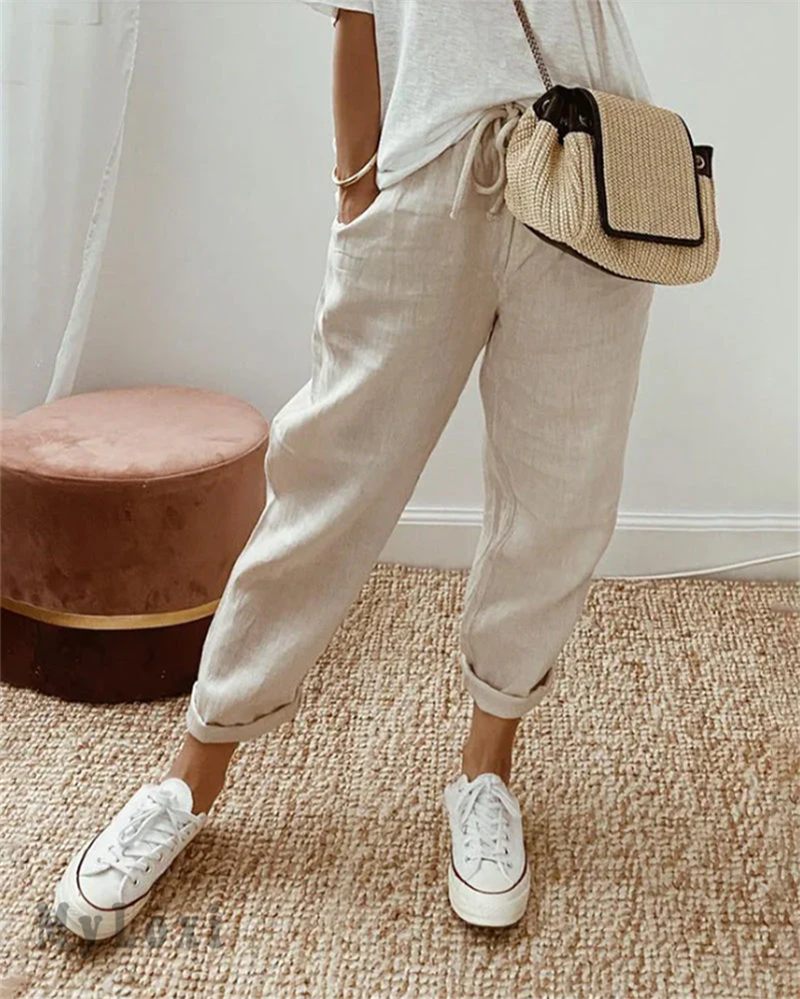 Beige casual pants on model with white sneakers, white top, woven bag, indoor setting with rug and plush seat.