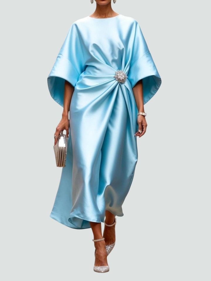 Elegant long satin dress in light blue with a floral brooch, styled with sparkly heels and a matching clutch, studio background.