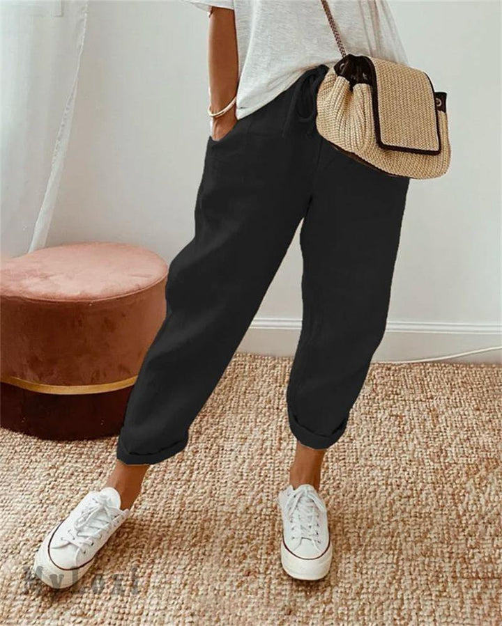 Black casual pants on model with white sneakers, white top, woven bag, indoor setting with rug and plush seat.