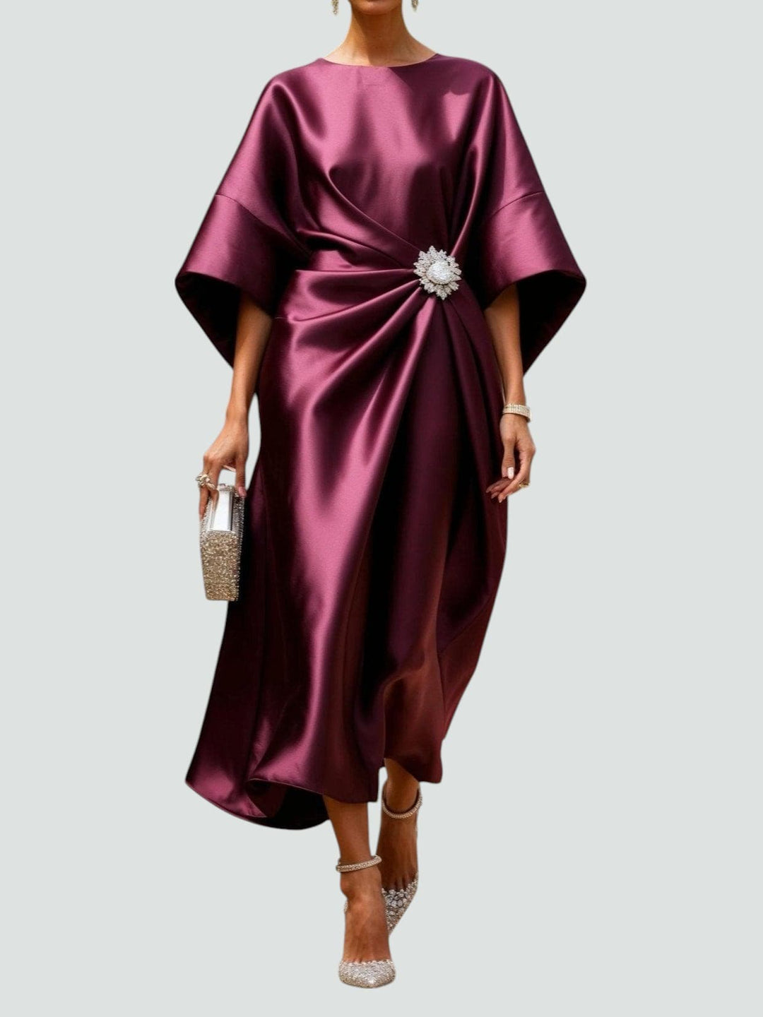 Elegant long satin dress in rich burgundy with a floral brooch, paired with sparkly heels and a matching clutch, studio background.