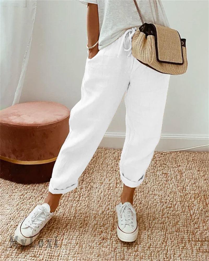 White casual pants on model with white sneakers, white top, woven bag, indoor setting with rug and plush seat.