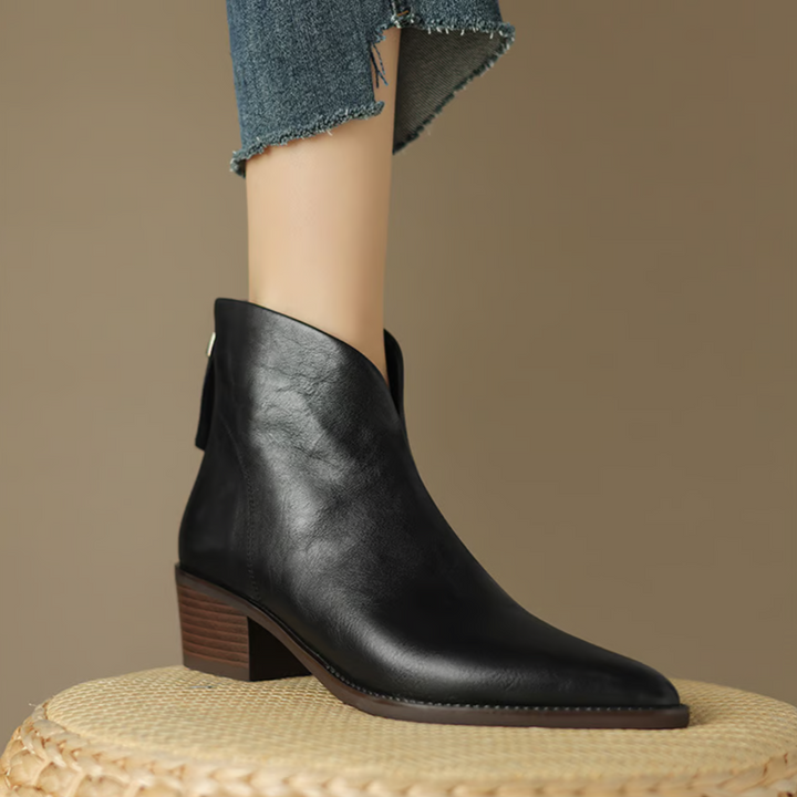 Black ankle boots with pointed toes, worn with frayed blue jeans in an indoor setting on a woven surface.