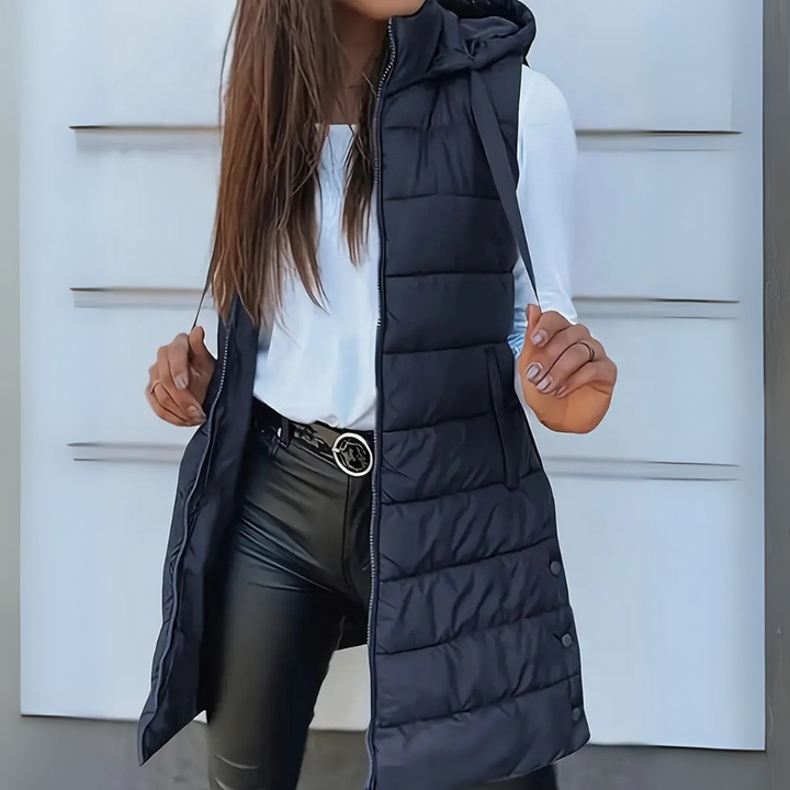 Black sleeveless puffer vest with hood worn over white top and black pants, outdoor setting.