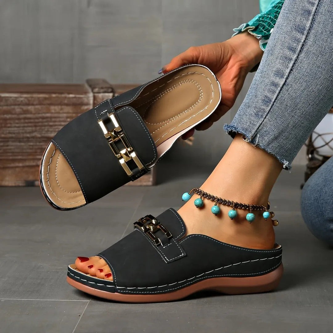 Black elegant comfort sandals with gold embellishments, paired with anklet, held against grey indoor background.