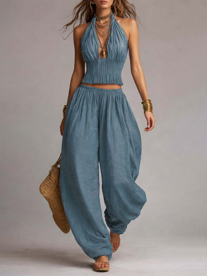 Blue two-piece set with a ruched top and billowy pants, accessorized with jewelry and straw bag. Studio background.