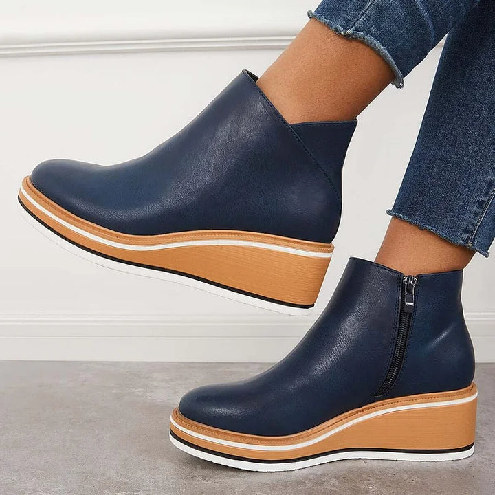 Side view of navy blue wedge boots with zipper, worn with jeans, against a white wall and gray floor.