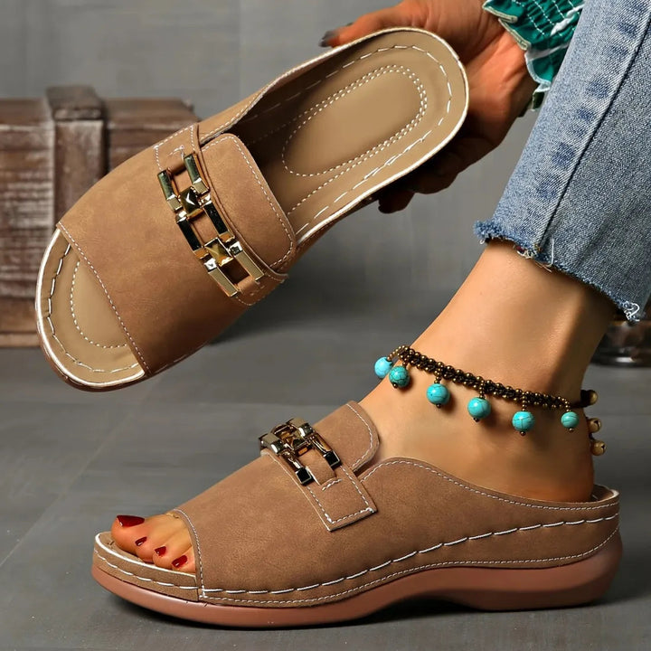 Brown comfort sandals with gold accents, worn with anklet, displayed indoors on grey floor with rustic decor.