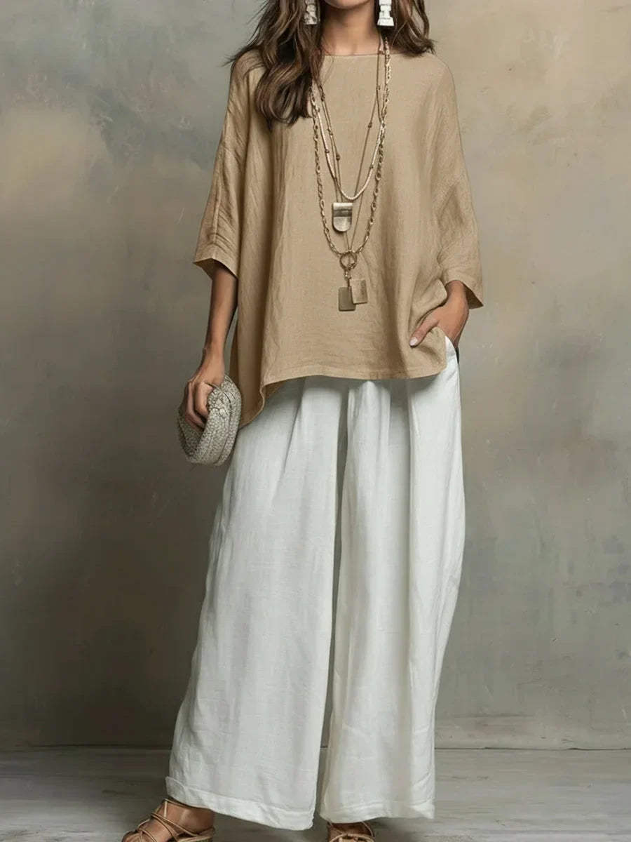 Woman in white and tan two-piece set, layered necklaces, holding textured clutch, studio backdrop.